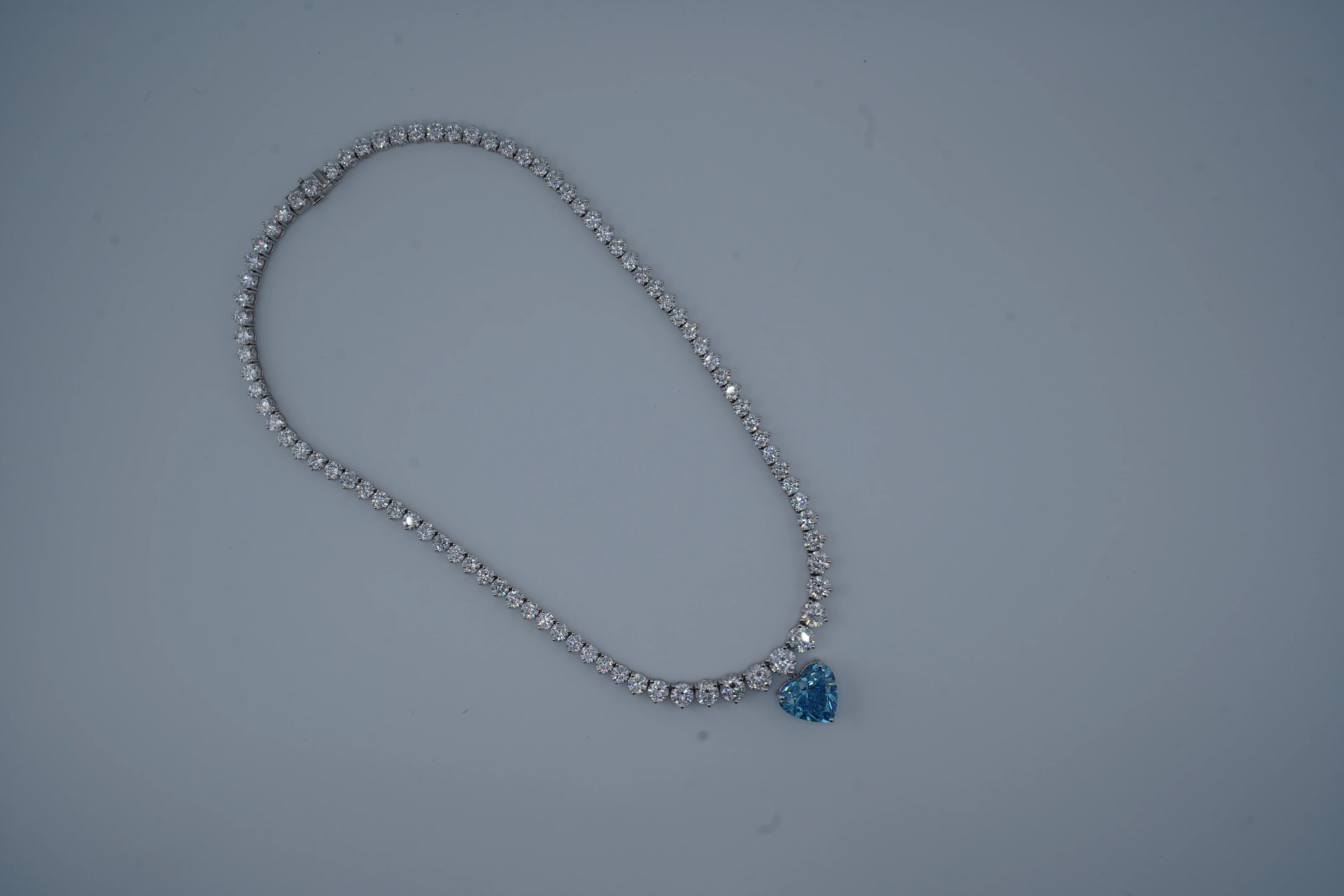 The raw studio frame: the blue heart tennis necklace lying in a loose, uneven loop on the blue-grey backdrop