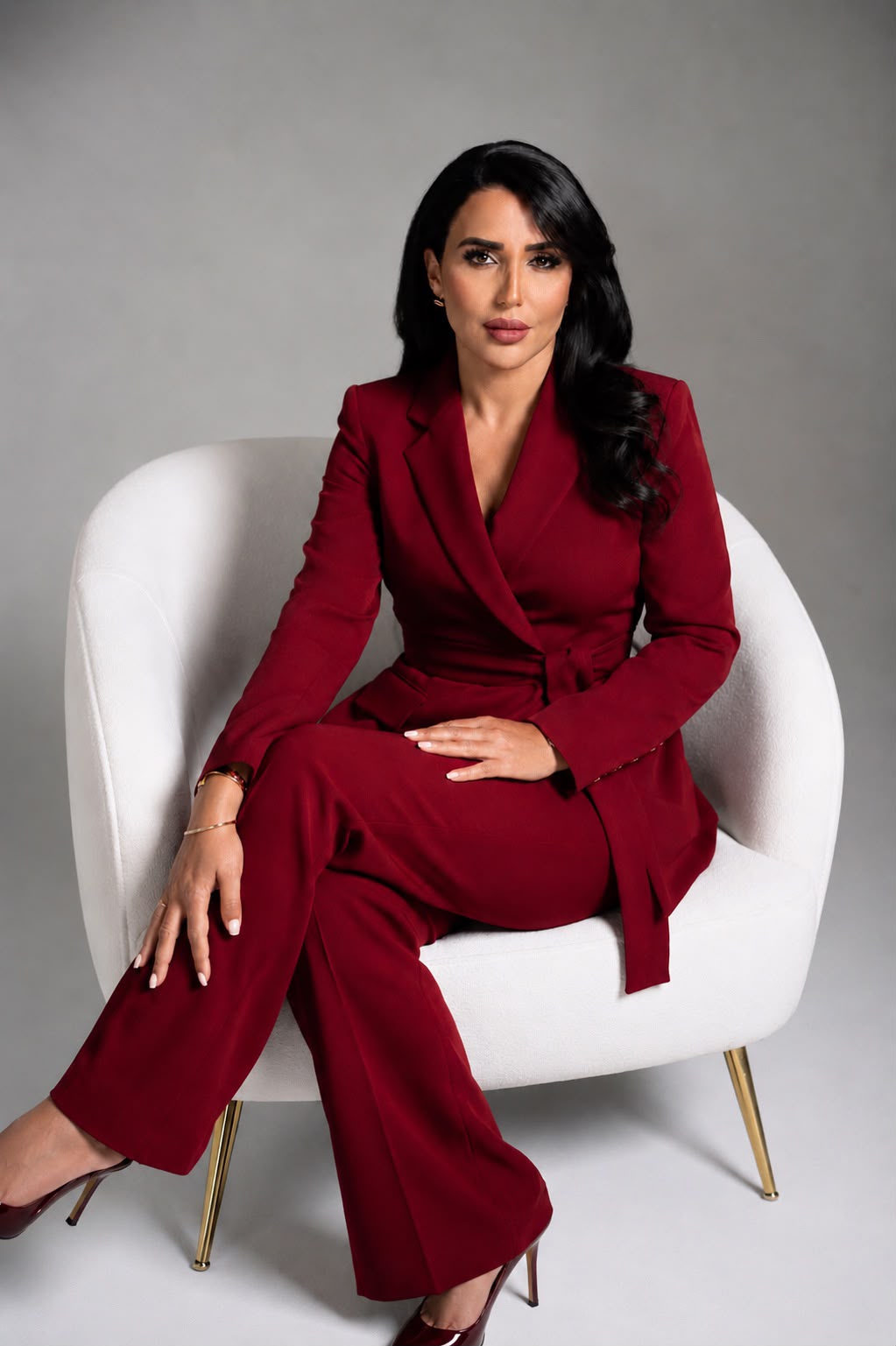 Sanaa Ouahmane in a crimson trouser suit, seated in a white armchair against a pale grey studio wall.