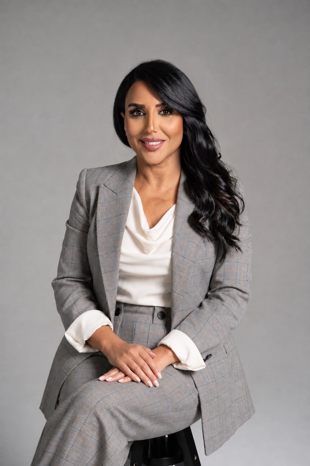 Sanaa Ouahmane in a grey check suit and white blouse, seated on a stool against a light grey studio wall.