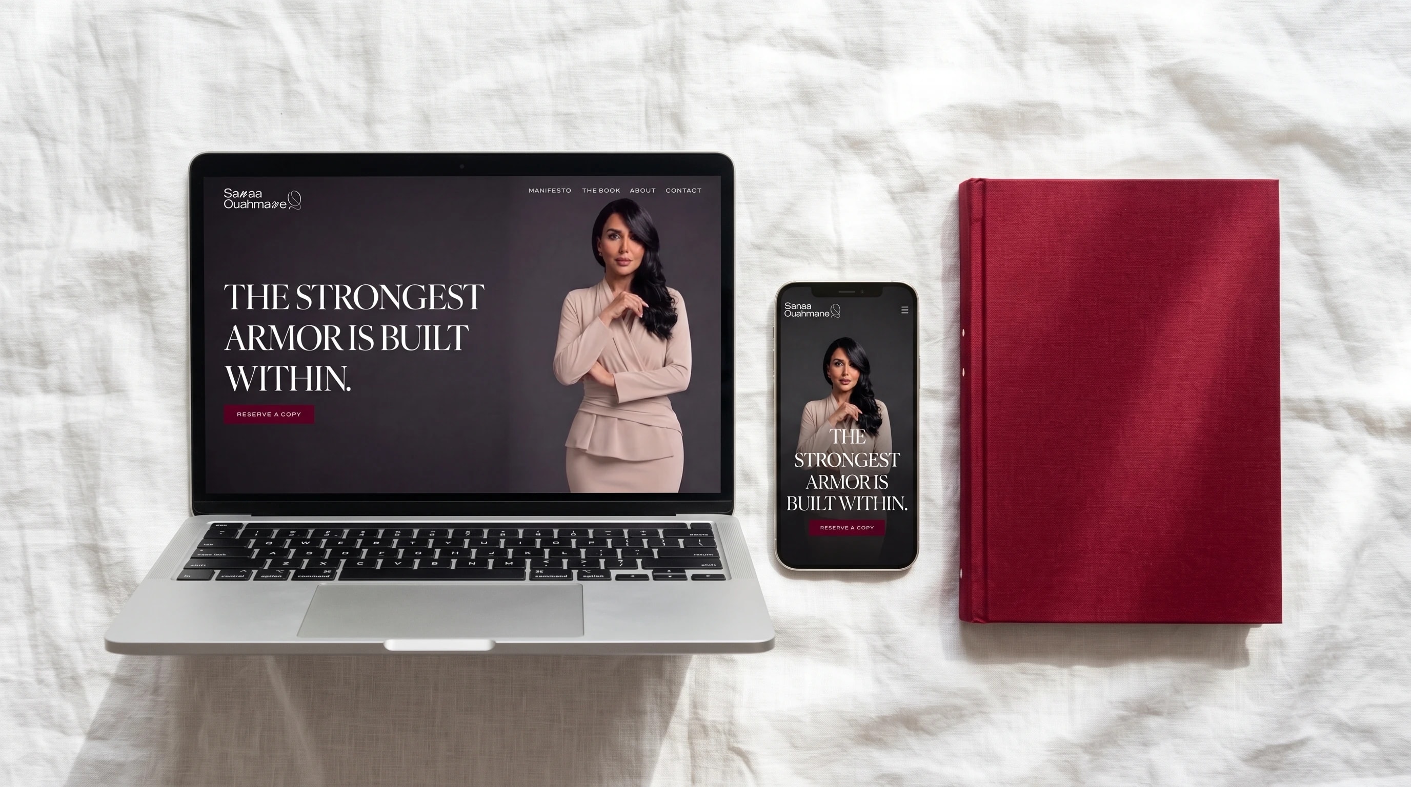 Sanaa Ouahmane's website on a laptop and a phone, on white linen beside a crimson cloth-bound book